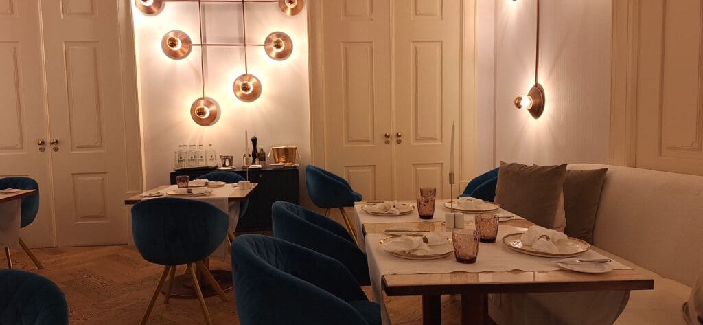 A cozy, elegantly decorated dining area with tables set for a meal. The tables have white tablecloths, gold-rimmed plates, and folded napkins. Blue cushioned chairs surround the tables. The walls are adorned with modern circular light fixtures, and there are decorative elements like a sideboard with bottles and a gold ice bucket. The lighting is warm, creating an inviting atmosphere.