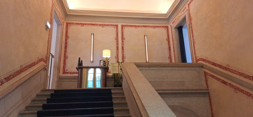 A staircase with a blue carpet leads up to a landing with a small desk and a lamp. The walls are decorated with ornate red patterns, and there is a stained glass window behind the desk. The ceiling is illuminated with soft lighting.