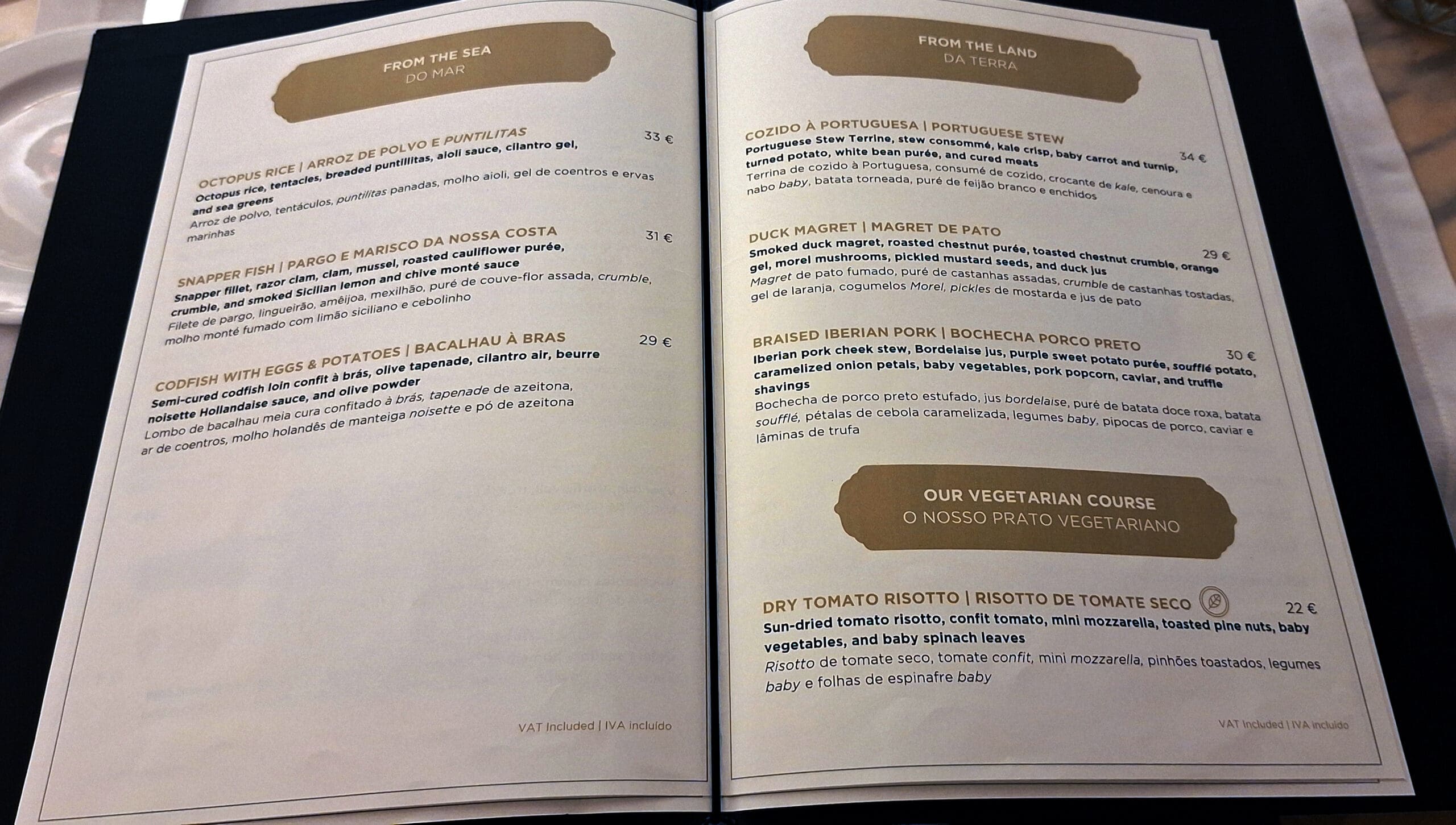 This image shows a restaurant menu with two pages. The left page is titled "From the Sea" and lists seafood dishes such as Octopus Rice, Snapper Fish, and Codfish with Eggs & Potatoes, with descriptions and prices in euros. The right page is titled "From the Land" and includes dishes like Portuguese Stew, Duck Magret, and Braised Iberian Pork. There is also a section for a vegetarian course featuring Dry Tomato Risotto. Each dish has a detailed description and price. The menu is bilingual, with descriptions in both English and Portuguese.