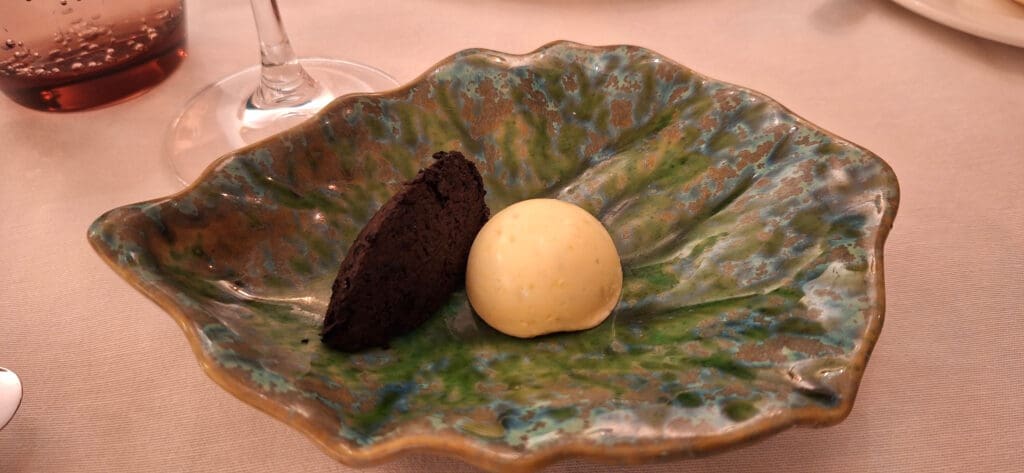 A decorative ceramic dish with a textured, multicolored glaze holds a small scoop of light yellow butter and a piece of dark bread. A wine glass is partially visible in the background.