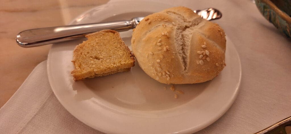 A white plate with a piece of sliced bread and a round bread roll topped with sesame seeds. A butter knife is placed on the plate, and the setting is on a table with a white tablecloth.