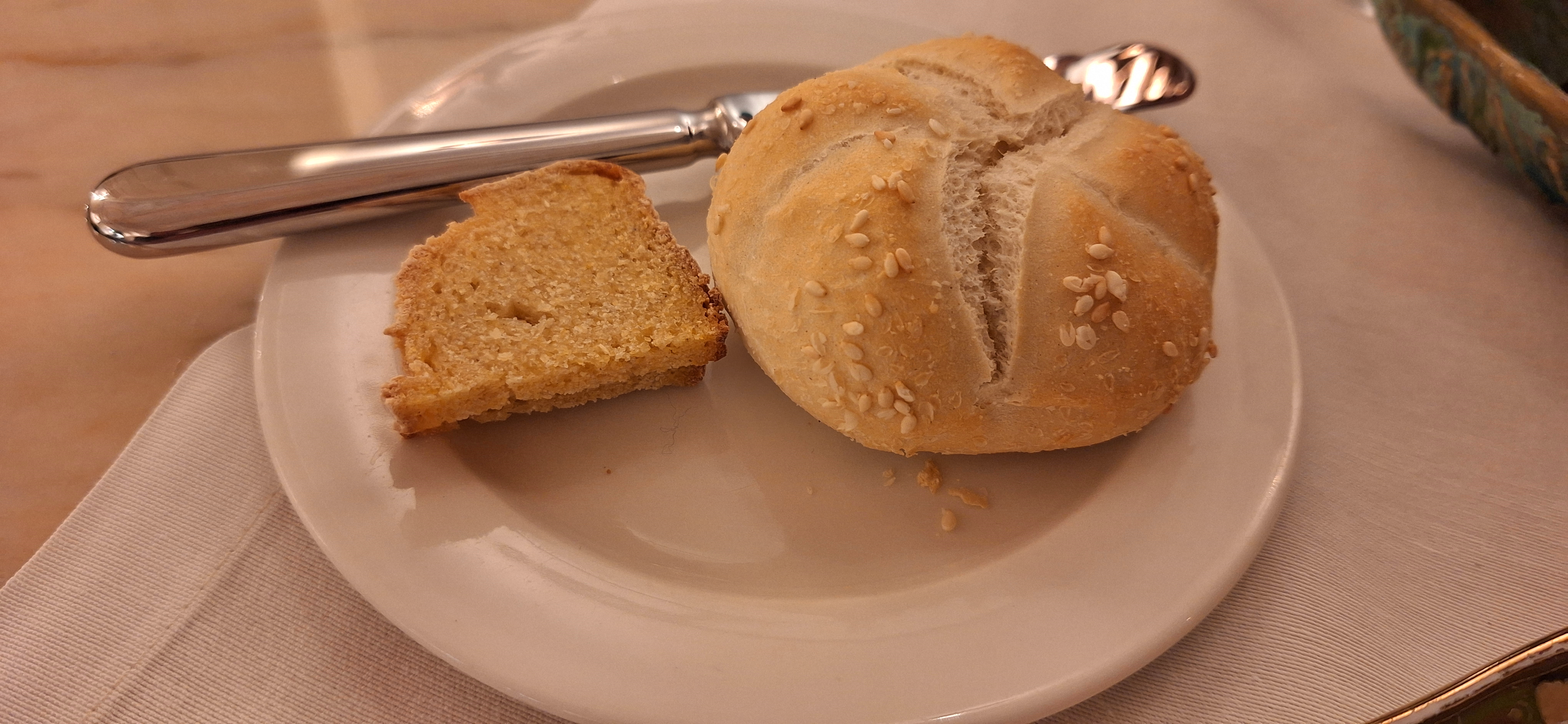 A white plate with a piece of sliced bread and a round bread roll topped with sesame seeds. A butter knife is placed on the plate, and the setting is on a table with a white tablecloth.