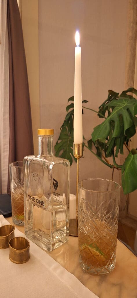 The image shows a table setting with a lit white candle in a gold holder, a clear glass bottle labeled "The One," and two crystal glasses containing a brown liquid with lime slices. There are also two gold napkin rings on a white tablecloth. In the background, there is a green plant with large leaves.