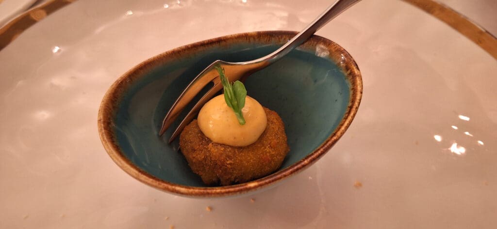 A small, round, breaded appetizer is placed in a blue, oval dish. It is topped with a dollop of creamy sauce and a small green garnish. A fork is resting on the edge of the dish. The dish is set on a white plate.