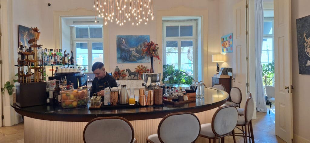 The image shows a stylish bar area with a bartender working behind a curved counter. The counter is adorned with various bottles, a basket of fruits, and drink-making equipment. Above the bar hangs an elegant chandelier with numerous small lights. The room has large windows, letting in natural light, and is decorated with paintings, plants, and autumn-themed floral arrangements. There are several chairs around the bar, and a lamp and books are visible on a side table.