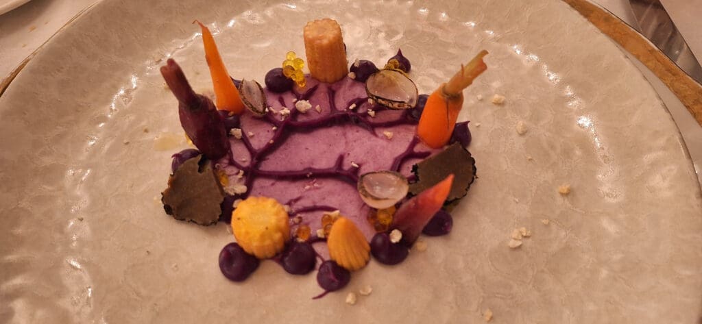 A gourmet dish is artfully presented on a textured plate. The dish features a purple sauce base with decorative elements including small carrots, corn, and other vegetables. There are also small dollops of purple puree, translucent slices, and yellow spherical garnishes, creating a visually appealing arrangement.