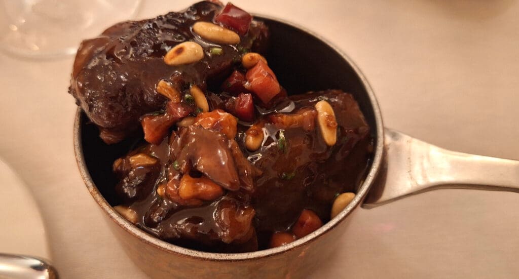 A close-up of a dish in a small metal pot, featuring braised meat pieces in a rich, dark sauce. The dish is garnished with pine nuts and small diced vegetables, possibly carrots and herbs. The pot has a handle and is placed on a light-colored surface.