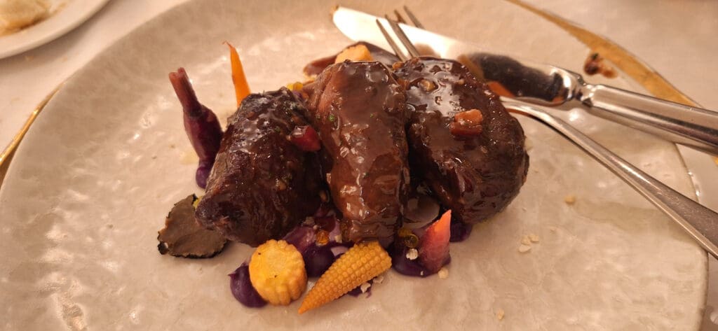 A gourmet dish featuring three pieces of braised meat covered in a glossy sauce, served on a plate with a variety of colorful vegetables, including baby corn, carrots, and purple cabbage. A knife and fork are placed beside the dish.