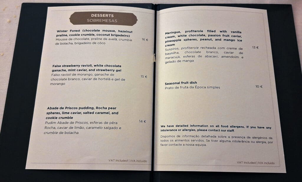 This image shows a dessert menu with items listed in both English and Portuguese. The menu includes: 1. **Winter Forest**: Chocolate mousse, hazelnut praline, cookie crumble, coconut brigadeiro - 16€. 2. **False Strawberry Ravioli**: White chocolate ganache, mint caviar, strawberry gel - 15€. 3. **Abade de Priscos Pudding**: Rocha pear spheres, lime caviar, salted caramel, cookie crumble - 14€. 4. **Meringue, Profiterole**: Filled with vanilla cream, white chocolate, passion fruit caviar, pineapple spheres, peanut, mango ice cream - 13€. 5. **Seasonal Fruit Dish**: 10€. The menu also mentions that detailed information on food allergens is available. Prices include VAT.