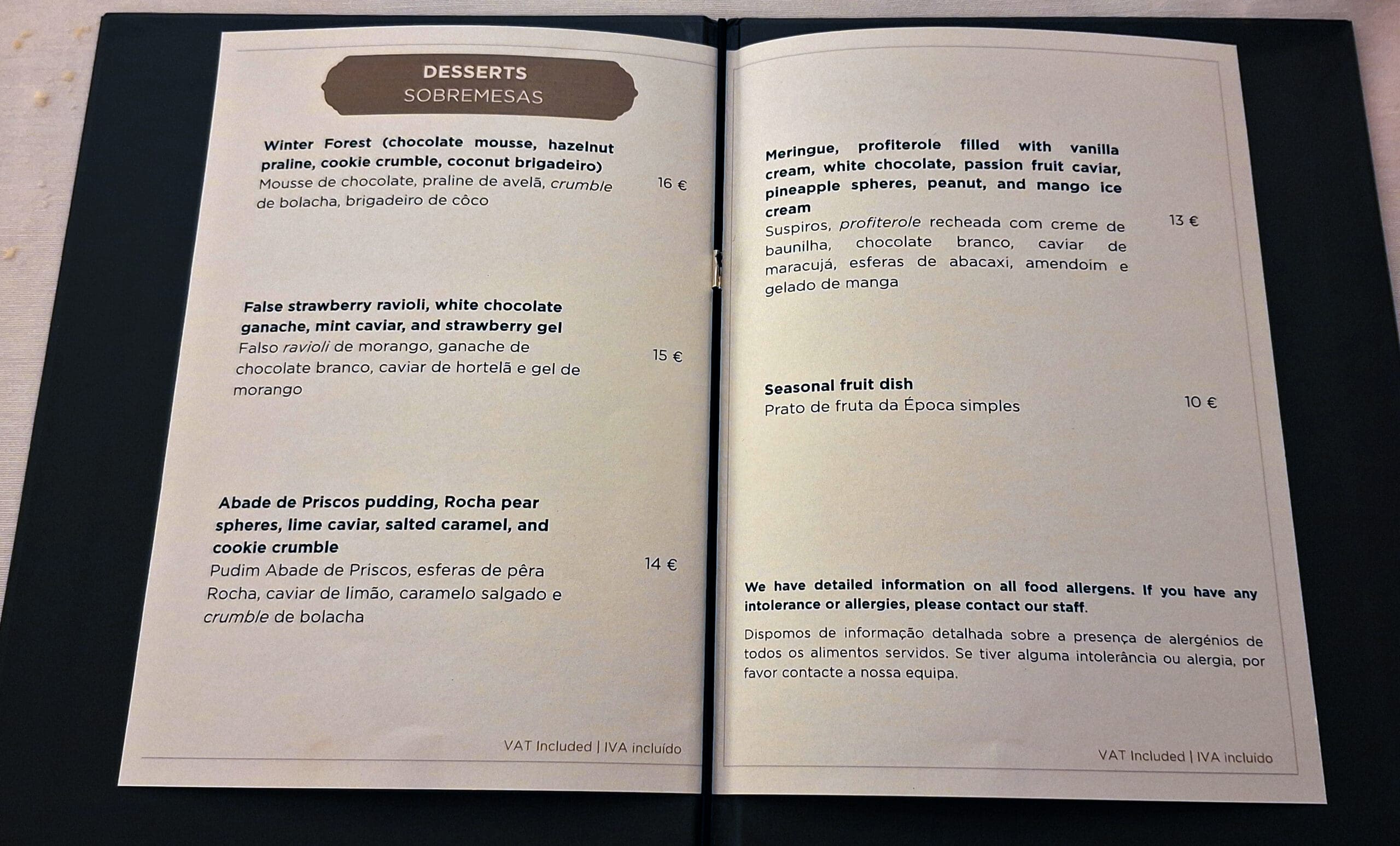 This image shows a dessert menu with items listed in both English and Portuguese. The menu includes:

1. **Winter Forest**: Chocolate mousse, hazelnut praline, cookie crumble, coconut brigadeiro - 16€.
2. **False Strawberry Ravioli**: White chocolate ganache, mint caviar, strawberry gel - 15€.
3. **Abade de Priscos Pudding**: Rocha pear spheres, lime caviar, salted caramel, cookie crumble - 14€.
4. **Meringue, Profiterole**: Filled with vanilla cream, white chocolate, passion fruit caviar, pineapple spheres, peanut, mango ice cream - 13€.
5. **Seasonal Fruit Dish**: 10€.

The menu also mentions that detailed information on food allergens is available. Prices include VAT.
