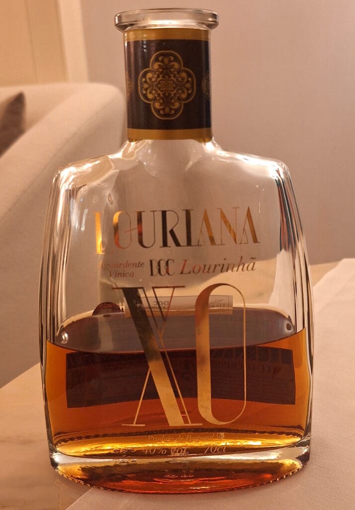 A bottle of Louriana XO Aguardente Vínica, a Portuguese brandy, is placed on a table. The bottle is clear with a decorative label featuring gold and black accents. The liquid inside is amber-colored. The background includes a soft-focus view of a couch.