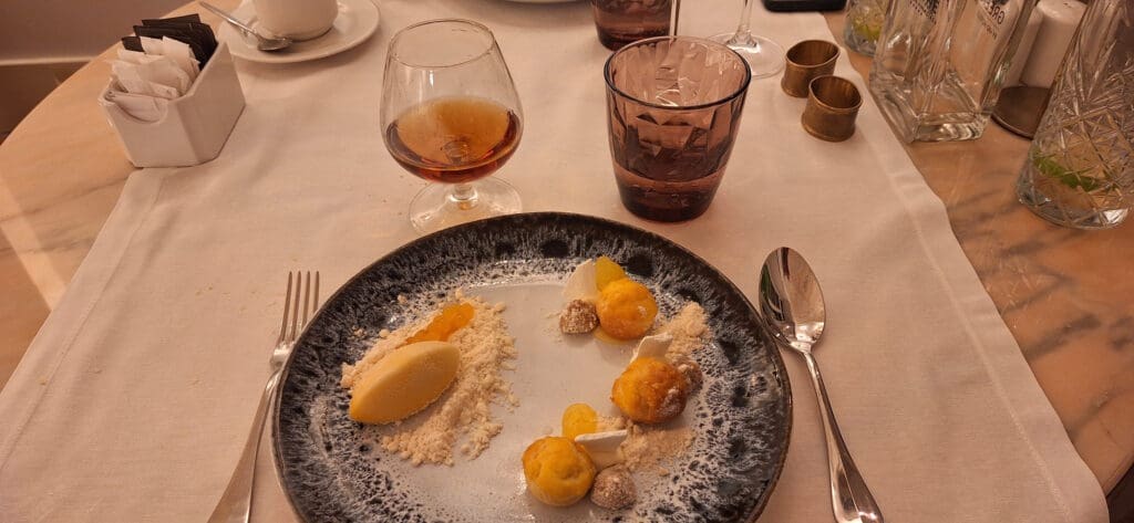 A dessert plate on a table with a variety of small pastries and a scoop of ice cream, garnished with crumbs and small fruit-like spheres. There is a glass of amber liquid, possibly a drink, next to the plate. The table is set with a fork, spoon, and a glass of water with a slice of lime. A cup with sugar packets and empty glasses are also visible.