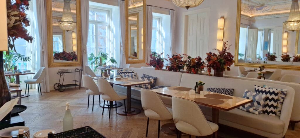 A stylish and elegant restaurant interior with white walls and large mirrors. The room features a long white cushioned bench with patterned pillows, surrounded by white chairs and wooden tables set with placemats and tableware. There are decorative plants and floral arrangements on the tables and along the bench. Large windows with white curtains allow natural light to fill the space, and a chandelier hangs from the ceiling, adding to the sophisticated ambiance.