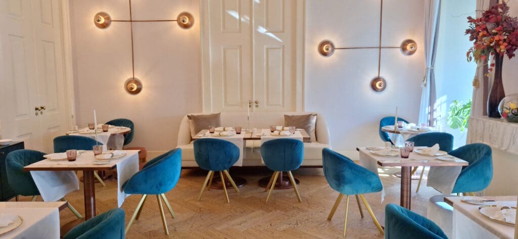 A stylish and elegant restaurant interior featuring several tables set for dining. The tables are adorned with white tablecloths, plates, glasses, and napkins. The chairs are upholstered in a rich blue fabric with gold legs. The walls are decorated with modern circular light fixtures, and there is a white cushioned bench along the back wall. A large vase with colorful flowers is placed on a marble surface to the right, and natural light streams in from a window, creating a warm and inviting atmosphere.