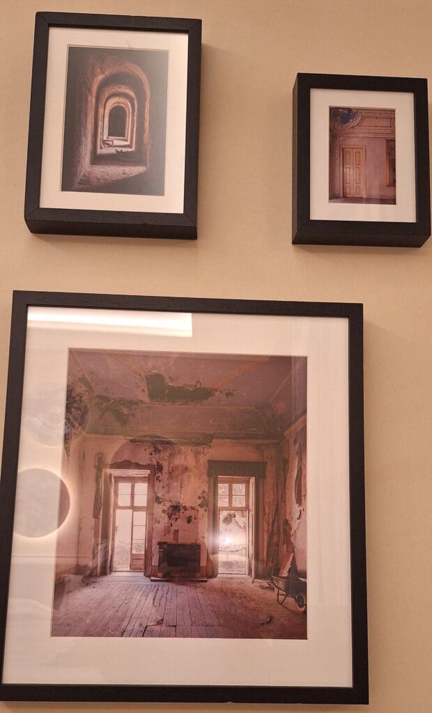 The image shows three framed photographs on a wall. The top left frame contains a photo of a long, arched hallway with a series of doorways. The top right frame features a photo of a room with a closed door and ornate ceiling details. The larger bottom frame displays a photo of an old, dilapidated room with peeling walls, two windows, and a wooden floor.