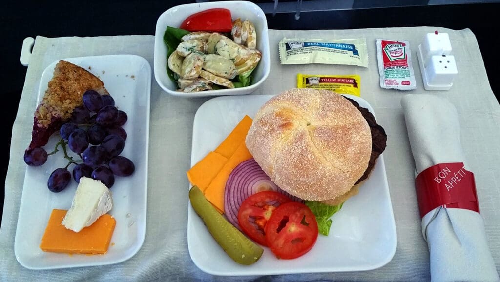 A meal is presented on a tray. It includes a hamburger with a bun, cheese slices, red onion, tomato slices, lettuce, and a pickle on a plate. There is a side salad with potatoes and a red pepper slice in a bowl. A dessert plate has a slice of pie, grapes, and two types of cheese. Condiment packets of mayonnaise, mustard, and ketchup are provided, along with salt and pepper shakers. A napkin is rolled with a red band labeled "Bon Appétit."