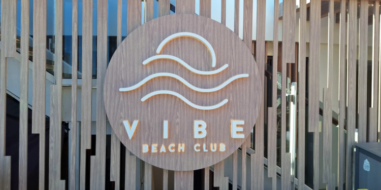The image shows a wooden sign with the words "VIBE BEACH CLUB" in white letters. Above the text, there is a design of a sun setting over waves. The sign is mounted on a wooden slatted background.