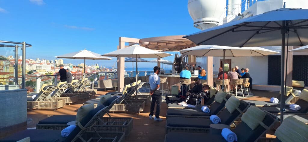 The image shows a rooftop deck with lounge chairs and umbrellas. People are relaxing on the chairs, and some are seated at a bar area. The deck overlooks a cityscape with colorful buildings under a clear blue sky. Towels are neatly placed on the lounge chairs, and a staff member is standing nearby.