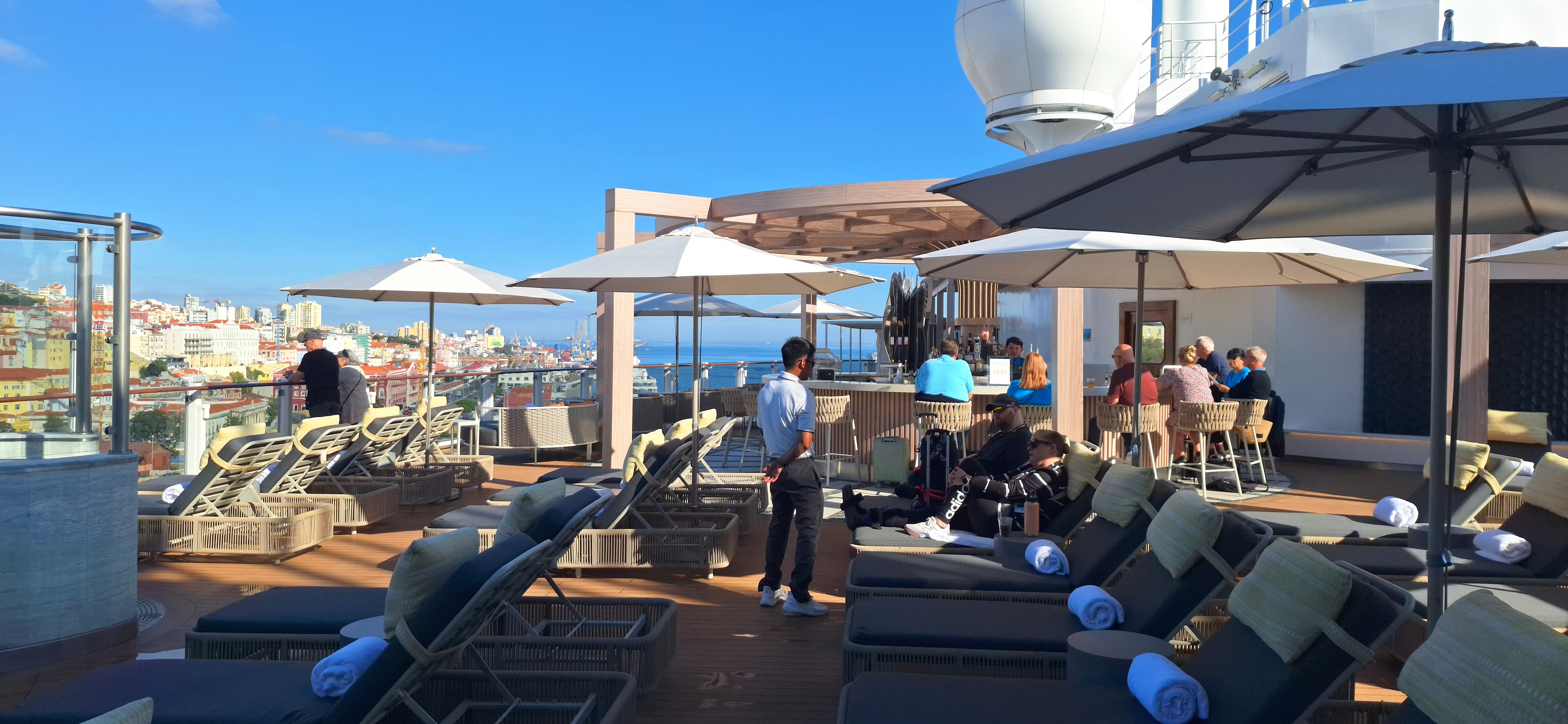 The image shows a rooftop deck with lounge chairs and umbrellas. People are relaxing on the chairs, and some are seated at a bar area. The deck overlooks a cityscape with colorful buildings under a clear blue sky. Towels are neatly placed on the lounge chairs, and a staff member is standing nearby.