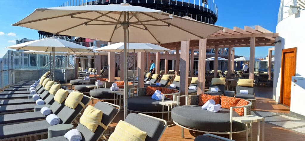 A luxurious outdoor lounge area on a cruise ship deck, featuring rows of cushioned sun loungers with rolled towels, large round daybeds with orange patterned pillows, and several large umbrellas providing shade. The area is surrounded by glass railings and has a wooden pergola structure. A few people are seen walking in the background.