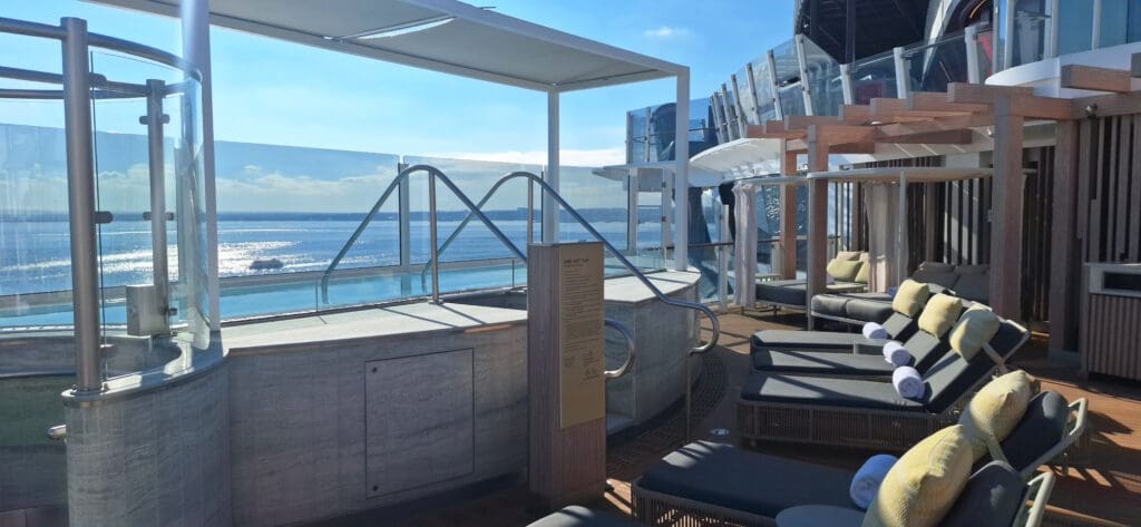 The image shows a luxurious outdoor deck area on a cruise ship. It features a hot tub with metal railings and a glass barrier overlooking the ocean. There are several cushioned lounge chairs with rolled towels and pillows under a wooden pergola. The sky is clear, and the sun is reflecting off the water, creating a serene and relaxing atmosphere.