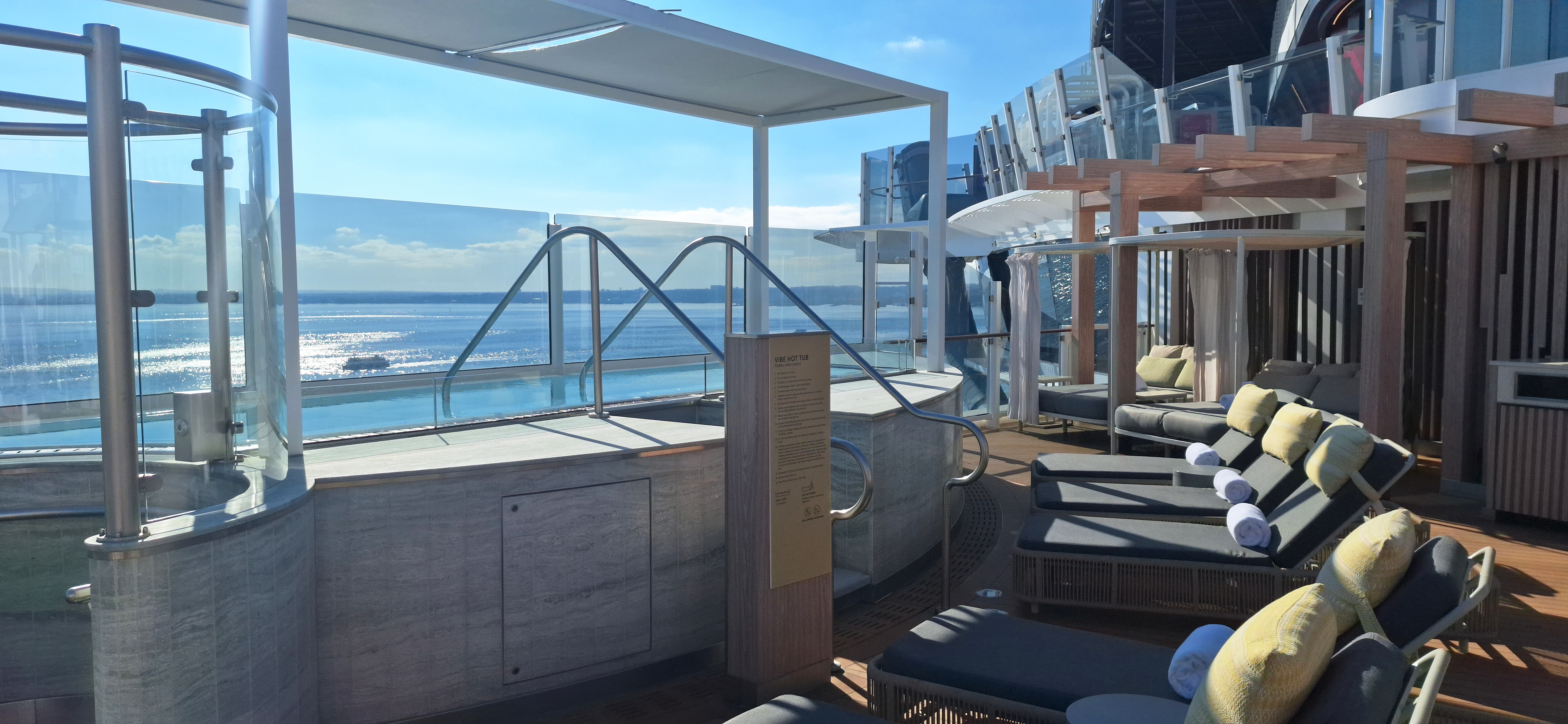 The image shows a luxurious outdoor deck area on a cruise ship. It features a hot tub with metal railings and a glass barrier overlooking the ocean. There are several cushioned lounge chairs with rolled towels and pillows under a wooden pergola. The sky is clear, and the sun is reflecting off the water, creating a serene and relaxing atmosphere.