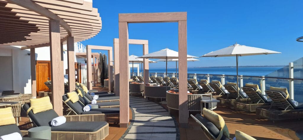 A luxurious outdoor deck area with several cushioned lounge chairs and umbrellas, set under a clear blue sky. The deck features wooden pergolas and overlooks a body of water, suggesting a relaxing, resort-like atmosphere.