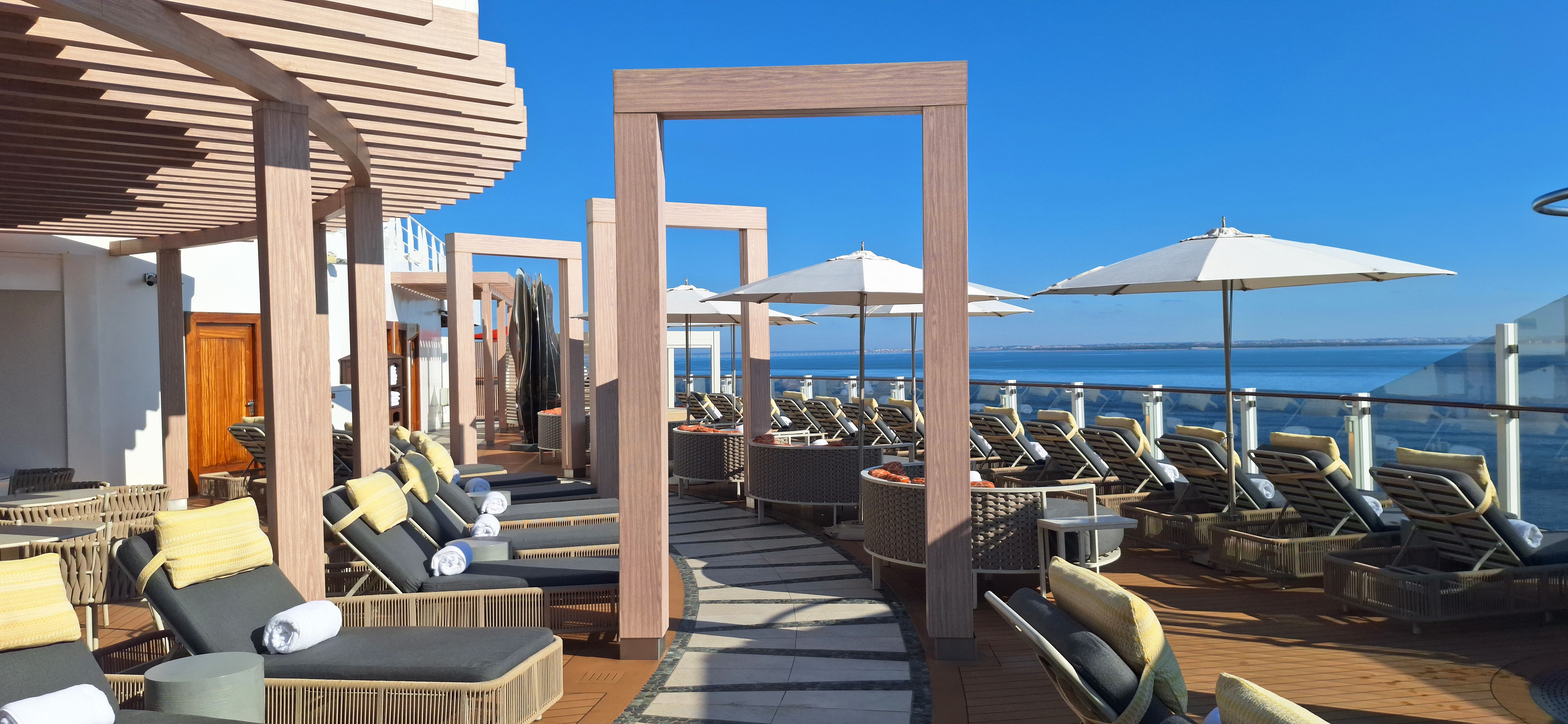 A luxurious outdoor deck area with several cushioned lounge chairs and umbrellas, set under a clear blue sky. The deck features wooden pergolas and overlooks a body of water, suggesting a relaxing, resort-like atmosphere.