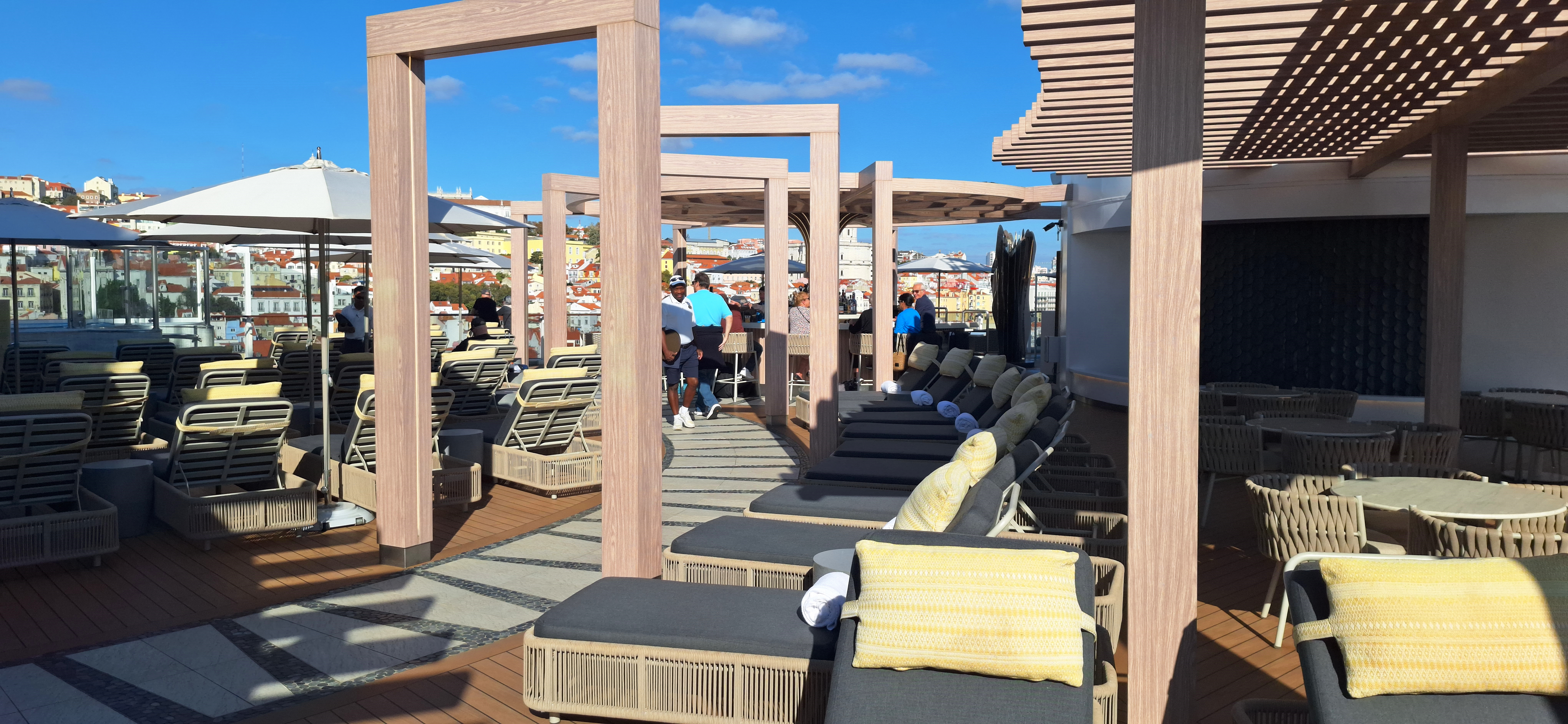 A rooftop terrace with lounge chairs and tables under wooden pergolas. There are people gathered near a bar area in the background. The terrace overlooks a cityscape with buildings under a clear blue sky.
