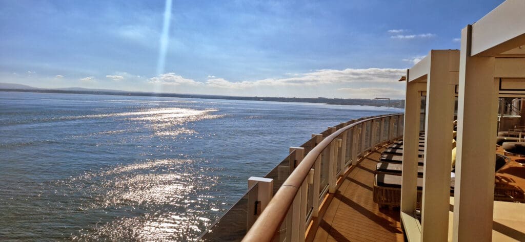 A view from a cruise ship deck overlooking a vast expanse of water under a clear blue sky. The sun reflects off the water, creating a sparkling effect. The deck has lounge chairs lined up along a glass railing, and part of the ship's structure is visible on the right. In the distance, there is a faint outline of land.