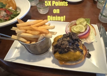 A plate of food featuring a cheeseburger topped with mushrooms, lettuce, tomato, pickles, and red onion, served with a side of French fries in a metal basket. The text "5X Points on Dining!" is displayed above the food. A salad is partially visible in the background.