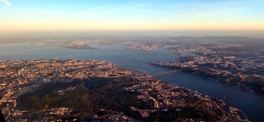 Aerial view of a cityscape at sunset, featuring a large body of water with a prominent red suspension bridge spanning across it. The city is densely packed with buildings, and there are areas of greenery visible. The sky is clear with a gradient from blue to orange near the horizon.