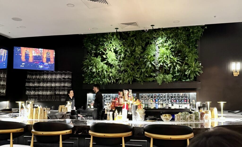 BLVD Steak in Calabasas, California. The image shows a modern bar with a sleek marble countertop and black and gold bar stools. Behind the bar, there is a lush green vertical garden on the wall. Two people, a man and a woman, are standing behind the bar, engaged in conversation. The bar is stocked with various bottles of alcohol and glassware. Above the bar, a television screen displays an image of a stage or performance. The lighting is warm and ambient, creating a cozy atmosphere.