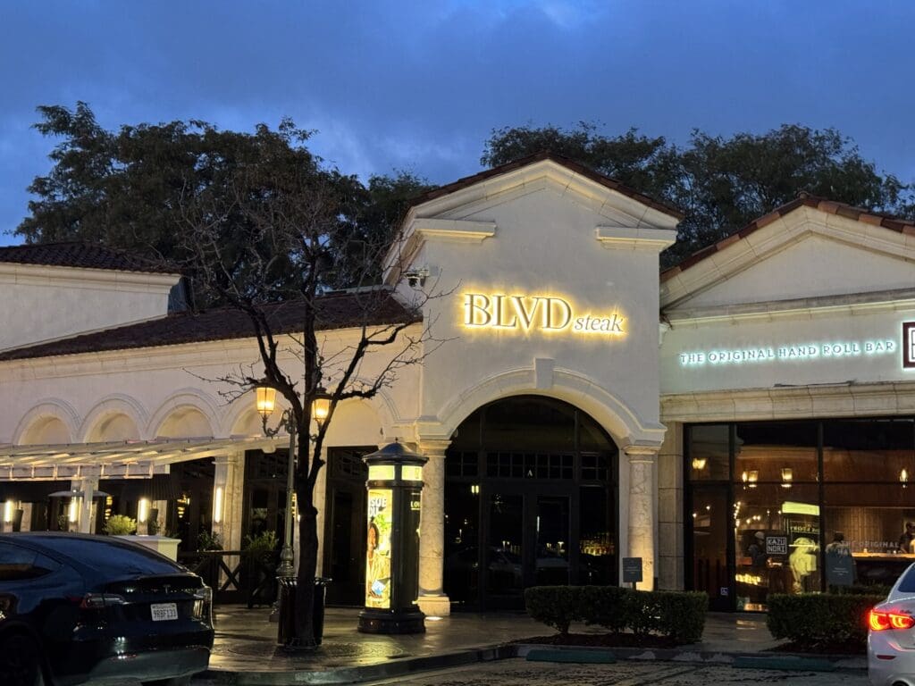 BLVD Steak in Calabasas, California. The image shows the exterior of a restaurant named "BLVD Steak" with illuminated signage. The building has an arched entrance and is flanked by another establishment with a sign that reads "The Original Hand Roll Bar." There are trees and street lamps in front of the building, and a parked car is visible in the foreground. The sky is overcast, suggesting it is either early evening or late afternoon.