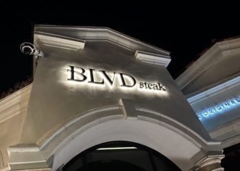 The image shows the exterior of a building at night with a sign that reads "BLVD steak" illuminated on the facade. The architecture features an arched design, and there is a security camera mounted on the corner of the building. Another partially visible sign in the background reads "ORIGINAL."