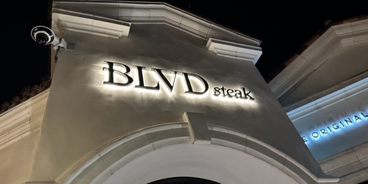 The image shows the exterior of a building at night with a sign that reads "BLVD steak" illuminated on the facade. The architecture features an arched design, and there is a security camera mounted on the corner of the building. Another partially visible sign in the background reads "ORIGINAL."