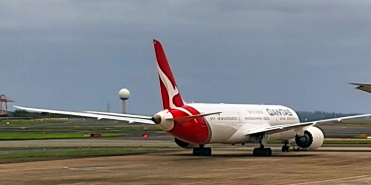 A Qantas airplane is on the tarmac at an airport. The aircraft is facing away from the camera, showing its tail with the distinctive red and white logo. The sky is overcast, and there are some airport structures and a radar tower visible in the background.