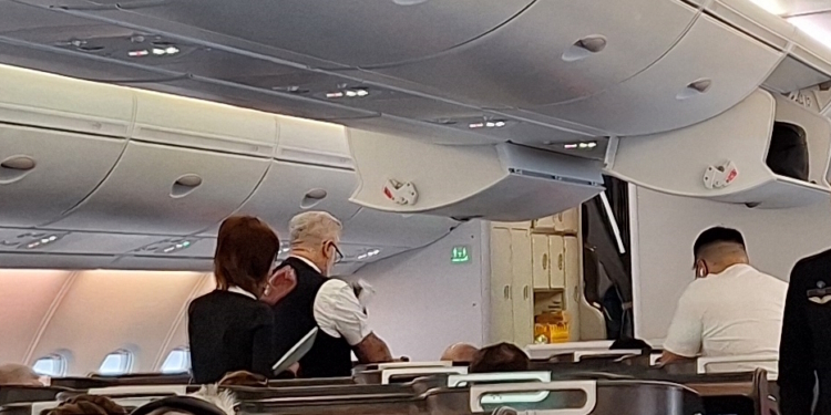 The image shows the interior of an airplane cabin. There are passengers seated, and two flight attendants are standing in the aisle. The overhead compartments are closed, and the cabin is well-lit. The flight attendants are wearing uniforms, and one is holding a tray. The cabin appears to be in a business or first-class section, with spacious seating.