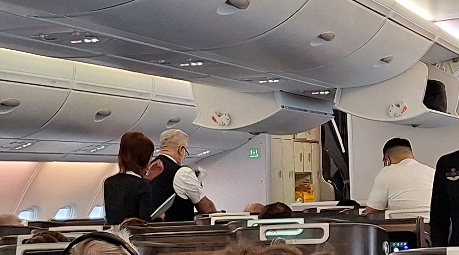 The image shows the interior of an airplane cabin. There are passengers seated, and two flight attendants are standing in the aisle. The overhead compartments are closed, and the cabin is well-lit. The flight attendants are wearing uniforms, and one is holding a tray. The cabin appears to be in a business or first-class section, with spacious seating.