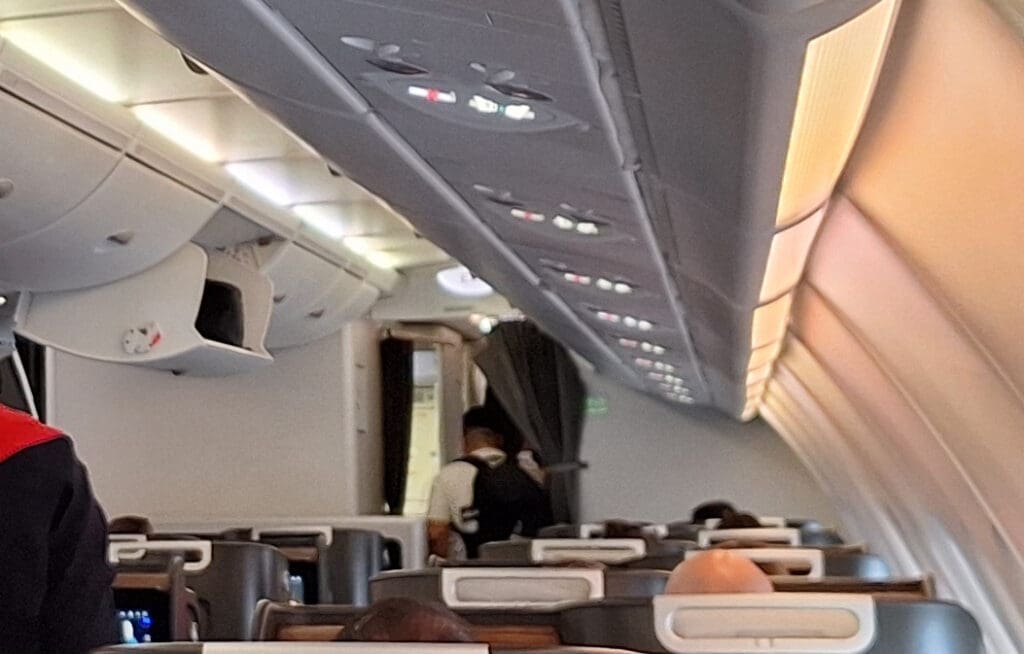 The image shows the interior of an airplane cabin. Overhead compartments are visible, some of which are open. Passengers are seated, and a person is standing near the exit door at the front of the cabin. The lighting is on, and the cabin appears to be in a business or first-class section.