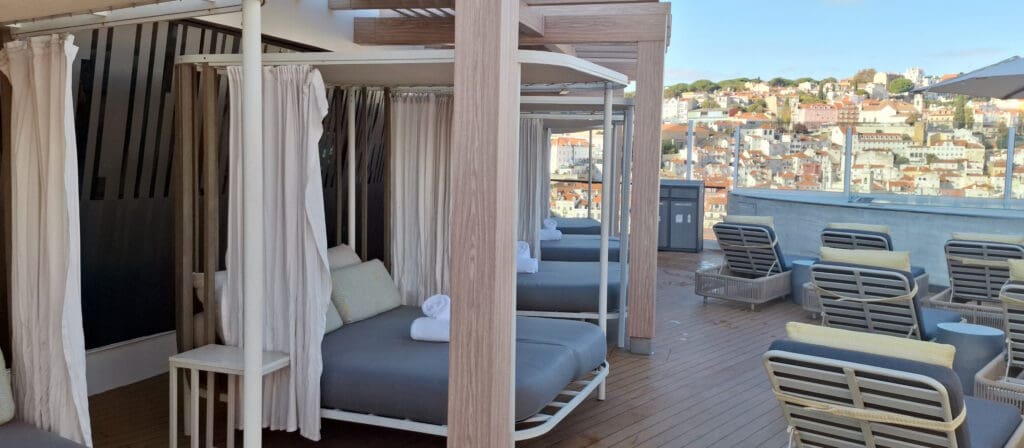 A rooftop terrace with several cabanas featuring white curtains and cushioned lounge beds. Rolled towels are placed on the beds. There are also wicker chairs with cushions arranged around small tables. The terrace overlooks a cityscape with numerous buildings and houses on a hillside. The sky is clear and blue.