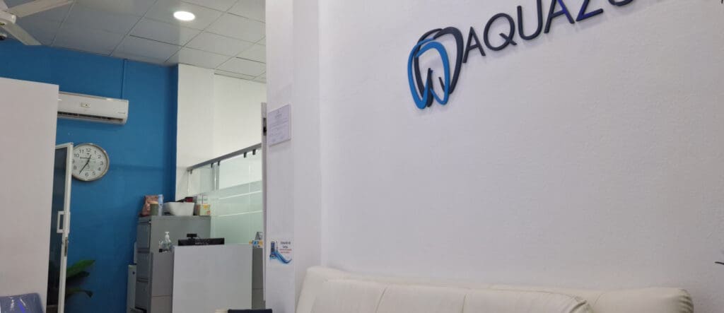 The image shows the interior of an office or clinic with a white and blue color scheme. On the right wall, there is a logo with the word "AQUAZO" and a stylized tooth design. A white couch is partially visible in the foreground. On the left, there is a blue wall with a clock, an air conditioning unit, and a glass door. A filing cabinet with various items on top is also visible. The ceiling has recessed lighting and a ceiling fan.