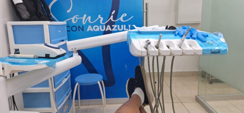 The image shows a dental clinic room with dental equipment. There is a dental chair with a person's legs visible, wearing black shoes and white socks. A dental instrument tray with various tools is covered with blue protective plastic. In the background, there is a blue wall with text and a storage unit with drawers. A stool is also visible near the chair.