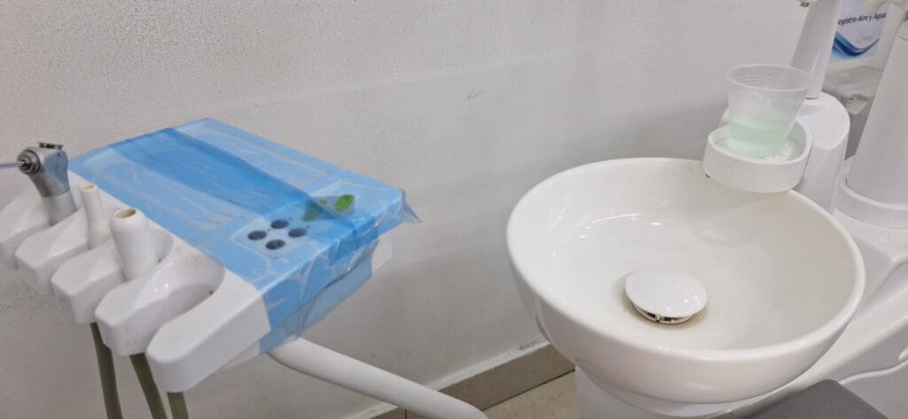 The image shows a dental office setup. On the left, there is a dental tool holder with various instruments, including a dental drill, covered with a blue protective film. On the right, there is a white dental sink with a small plastic cup containing a green liquid placed on a holder. The background is a plain white wall.