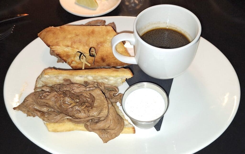 French dip at BLVD Steak in Calabasas, California. A plate with a roast beef sandwich on toasted bread, secured with toothpicks. Next to the sandwich is a small cup of creamy dipping sauce. A white cup of black coffee is placed on the plate, resting on a black napkin. In the background, there is a small plate with a lemon wedge.