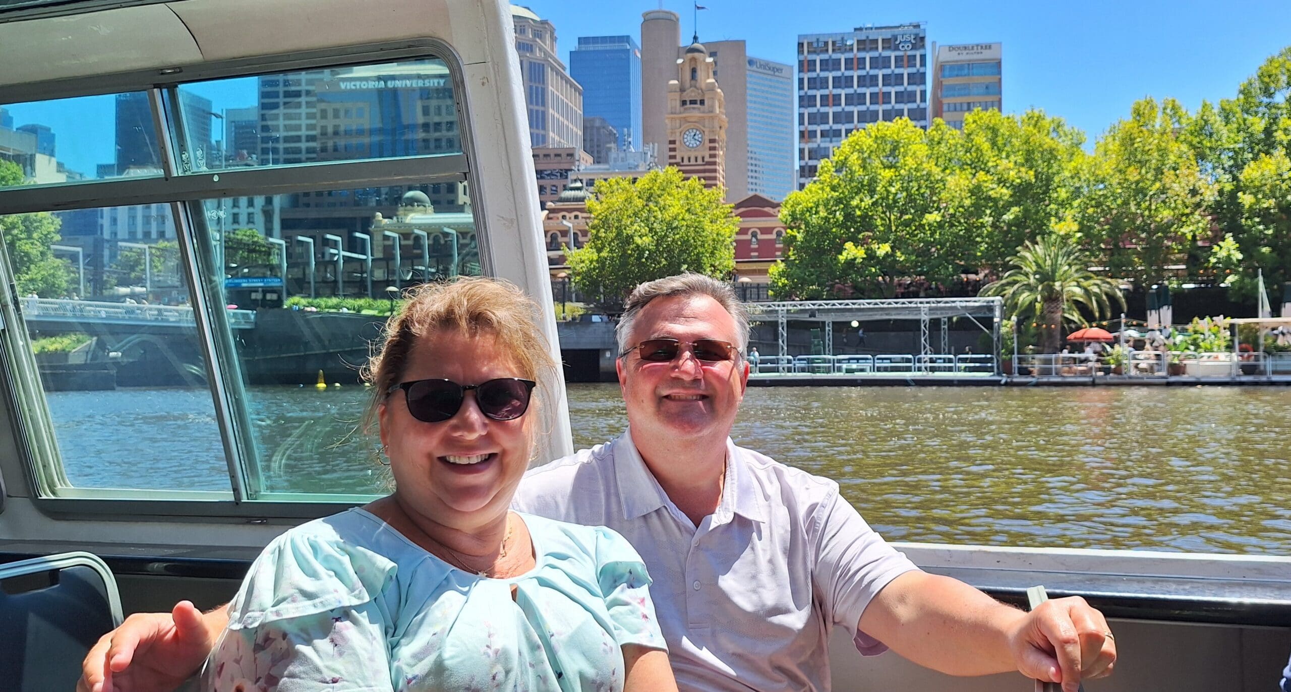 A man and a woman are sitting on a boat, smiling at the camera. They are both wearing sunglasses. In the background, there is a river with trees and buildings, including a clock tower. The sky is clear and blue.
