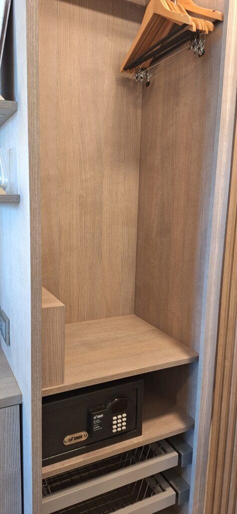 El interior de un armario de madera con varias perchas de madera sobre un riel en la parte superior. Debajo de las perchas hay un estante con una caja fuerte digital. Debajo del armario se encuentran dos cestas de alambre que se pueden extraer.