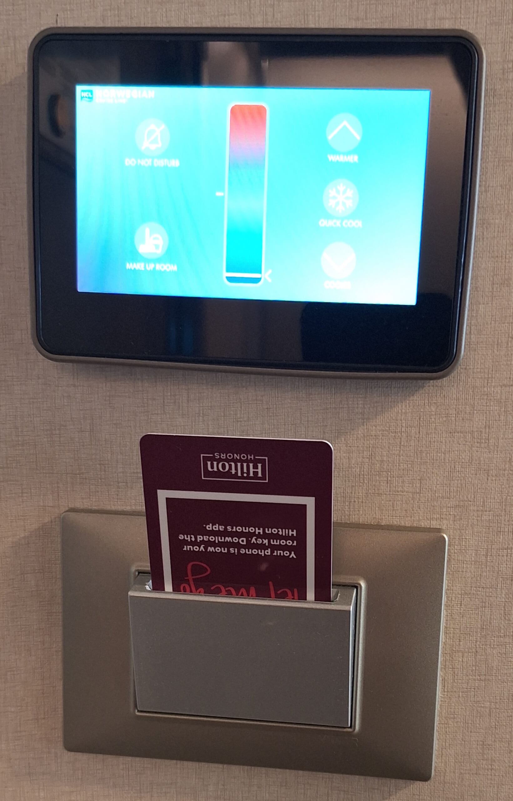 The image shows a hotel room control panel mounted on a wall. The panel has a digital display with options such as "Do Not Disturb," "Make Up Room," "Warmer," and "Quick Cool." Below the panel, there is a card slot with a hotel key card inserted, which has the Hilton Honors logo on it.