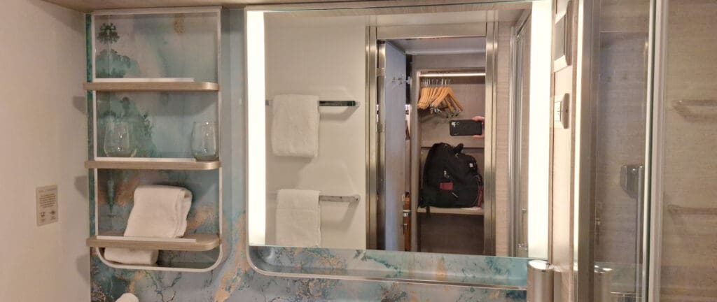 La foto muestra un baño moderno con un gran espejo y bordes iluminados. A la izquierda hay estanterías con toallas blancas dobladas y cristal. La pared presenta un patrón decorativo azul y beige. En el reflejo del espejo hay un armario con perchas y una mochila.