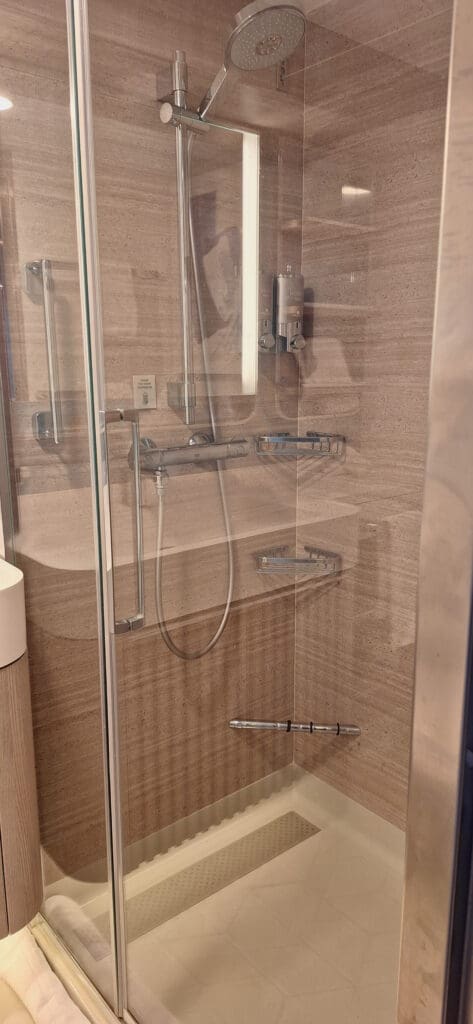 Esta foto muestra una cabina de ducha moderna con puertas de vidrio y manija cromada. En el interior hay un cabezal de ducha tipo lluvia y un accesorio de ducha de mano. Las paredes están revestidas con un material beige parecido a la piedra. Hay dos dispensadores de pared y un estante metálico para artículos de baño. El suelo de la ducha es blanco con cinta adhesiva.